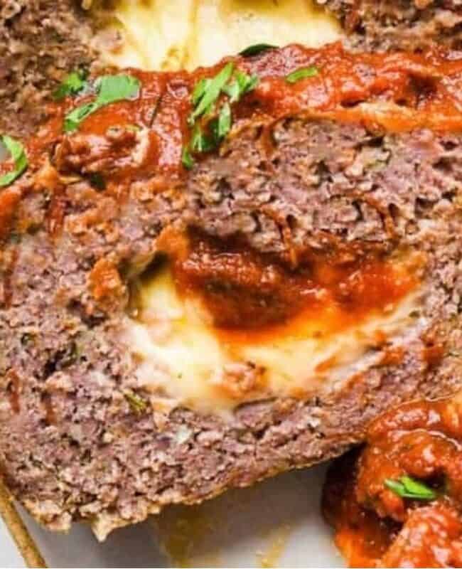 Close-up of a slice of meatloaf stuffed with melted cheese and topped with tomato sauce and herbs. Parsley garnish is visible on the side.