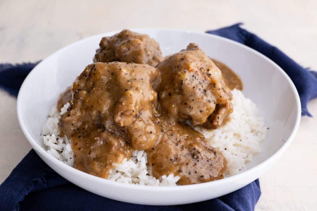 A white bowl filled with white rice topped with several pieces of meat coated in thick brown gravy. The bowl is placed on a dark blue cloth.