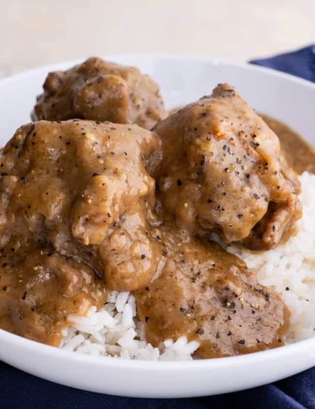 A white bowl filled with white rice topped with several pieces of meat coated in thick brown gravy. The bowl is placed on a dark blue cloth.