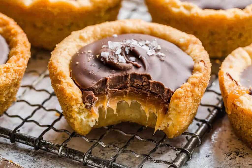 A close-up of a chocolate caramel tart with a bite taken out, revealing the gooey caramel center. The tart has a golden-brown crust and is topped with dark chocolate and a sprinkle of sea salt. Other similar tarts are visible in the background.