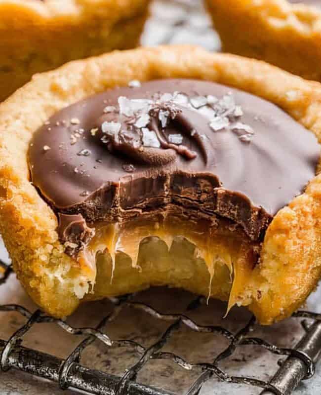 A close-up of a chocolate caramel tart with a bite taken out, revealing the gooey caramel center. The tart has a golden-brown crust and is topped with dark chocolate and a sprinkle of sea salt. Other similar tarts are visible in the background.