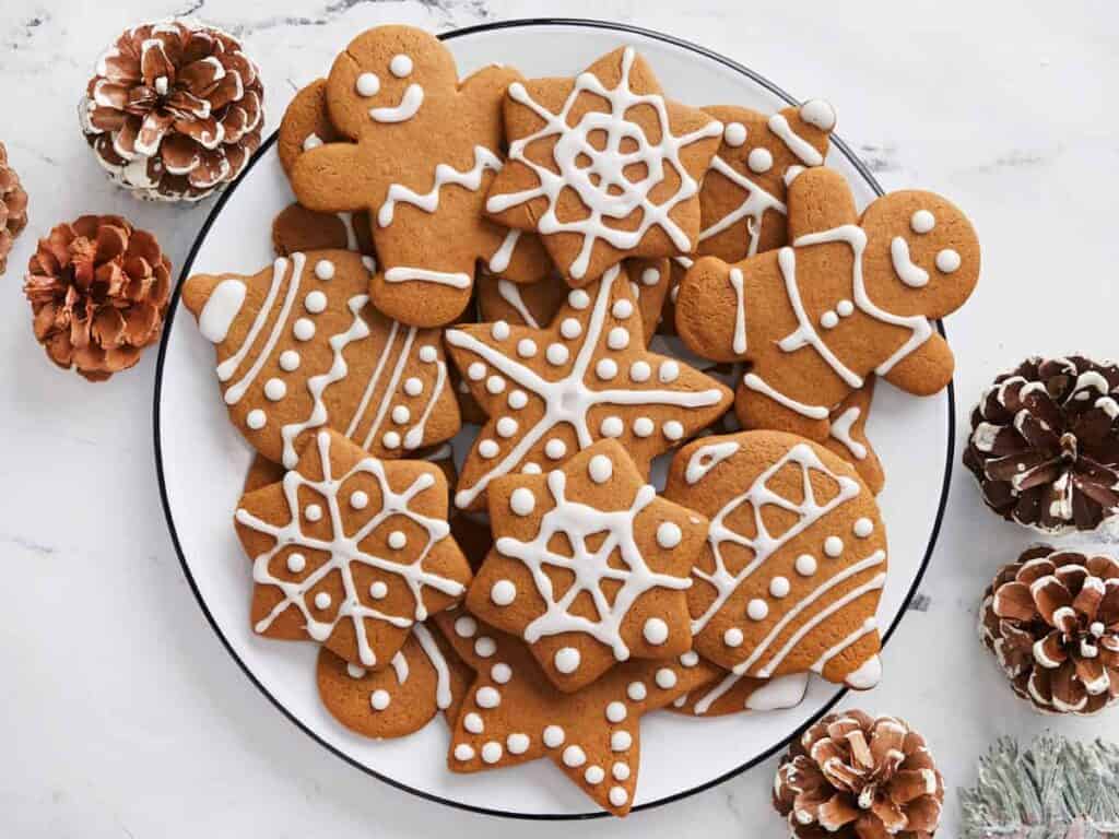 A plate of gingerbread cookies decorated with white icing, featuring shapes like stars, snowflakes, gingerbread people, and trees. The cookies are surrounded by pinecones on a marble surface.