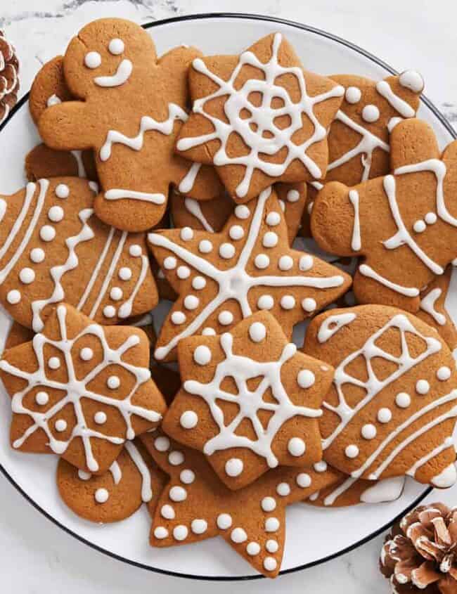 A plate of gingerbread cookies decorated with white icing, featuring shapes like stars, snowflakes, gingerbread people, and trees. The cookies are surrounded by pinecones on a marble surface.