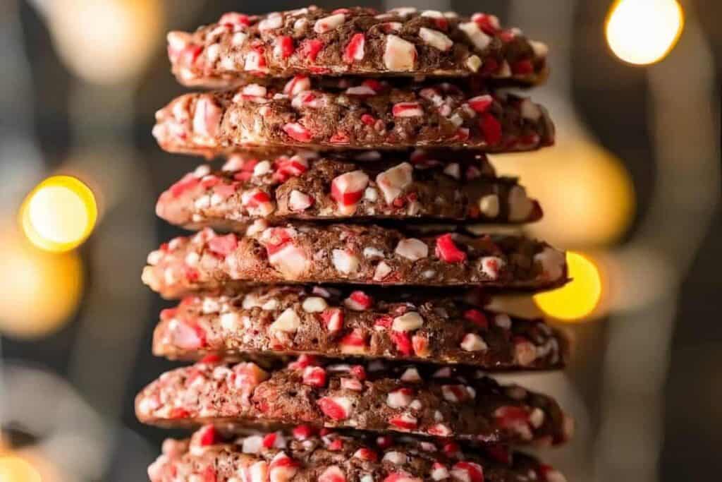 A stack of chocolate cookies studded with red and white peppermint pieces. The cookies are piled high against a soft-focus background of warm yellow lights, creating a festive and cozy atmosphere.