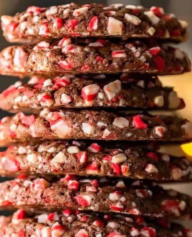 A stack of chocolate cookies studded with red and white peppermint pieces. The cookies are piled high against a soft-focus background of warm yellow lights, creating a festive and cozy atmosphere.