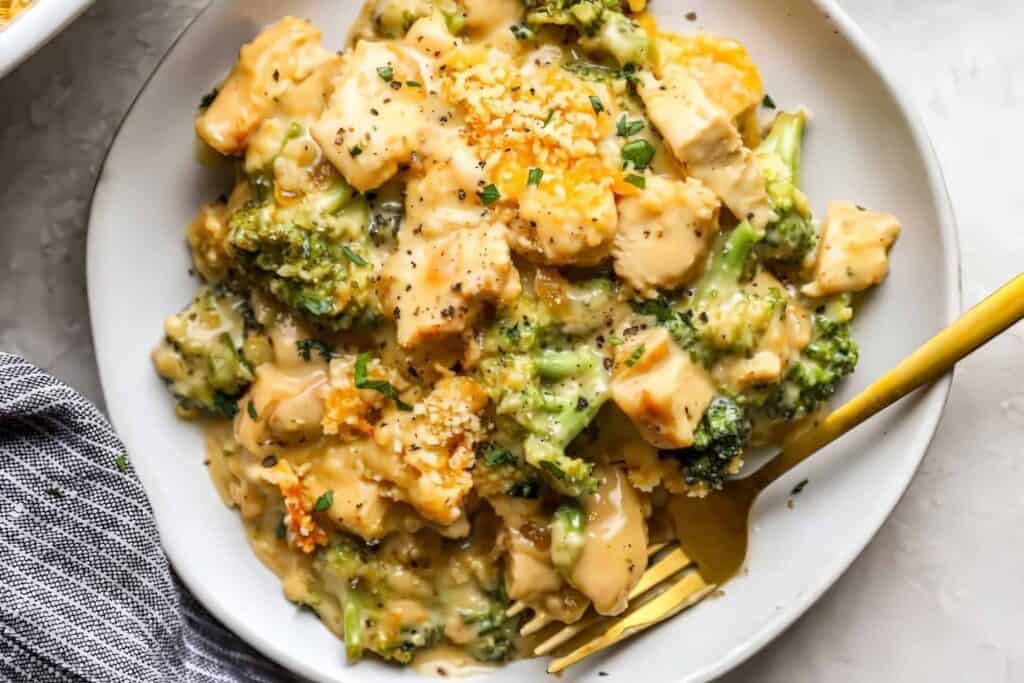 A bowl of creamy pasta with broccoli and chunks of chicken, showcasing one of the delightful chicken recipes. Topped with grated cheese and black pepper, a fork rests nearby. A striped cloth is partially visible at the bottom left corner, inviting you to savor every bite.