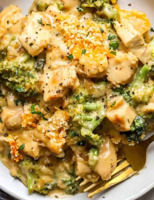 A bowl of creamy pasta with broccoli and chunks of chicken, showcasing one of the delightful chicken recipes. Topped with grated cheese and black pepper, a fork rests nearby. A striped cloth is partially visible at the bottom left corner, inviting you to savor every bite.