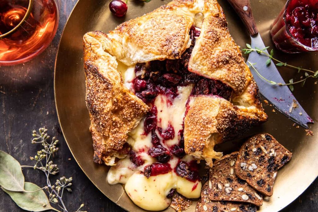 A baked brie en croûte with cranberry filling, partially sliced open, sits on a gold plate as the perfect Thanksgiving appetizer. Surrounding it are crackers, a knife, and a jar of cranberry sauce. A glass of rosé and scattered herbs complete the rustic setting.