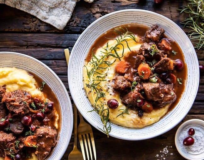 Two bowls of beef stew with carrots and cranberries, served over creamy mashed potatoes and garnished with rosemary, grace the rustic wooden table. A gold fork and napkin sit beside them—perfect for those eager to explore new stew recipes.