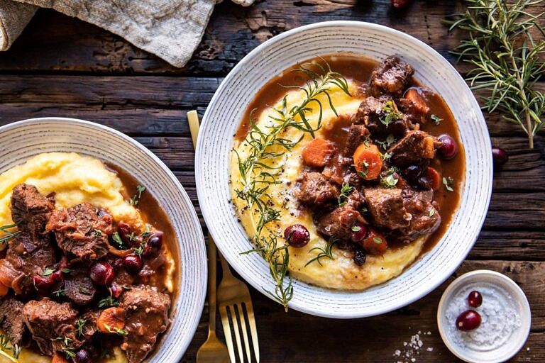 Two bowls of beef stew with carrots and cranberries, served over creamy mashed potatoes and garnished with rosemary, grace the rustic wooden table. A gold fork and napkin sit beside them—perfect for those eager to explore new stew recipes.