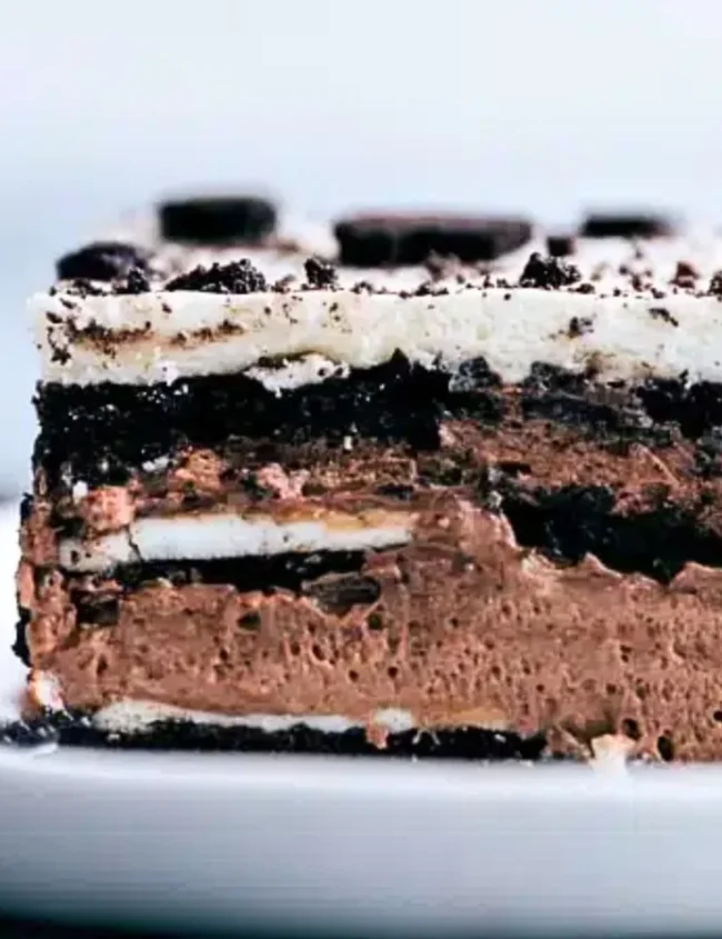 A slice of Oreo lasagna sits on a white plate, showcasing layers from the finest whipped cream recipes, chocolate mousse, and Oreo cookies. It's topped with crumbled Oreos, while a fork rests invitingly beside the dessert.