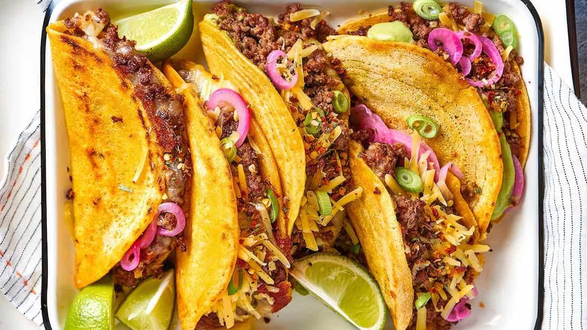 A tray of four delicious tacos features seasoned ground beef from beloved Ground Beef Recipes, topped with shredded cheese, sliced green onions, and pickled red onions. Lime wedges are scattered around the tacos for garnish and added flavor.