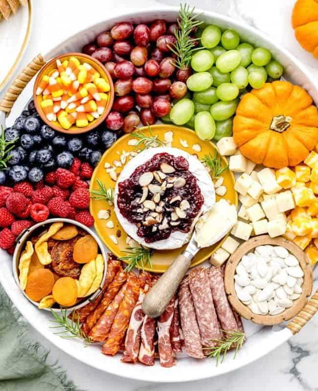 A vibrant charcuterie board perfect for Thanksgiving appetizers features a cheese wheel topped with cranberries and almonds, surrounded by grapes, berries, apricots, assorted cheeses, cured meats, crackers, and small pumpkins. Nearby are plates with sliced bread and golden cutlery.