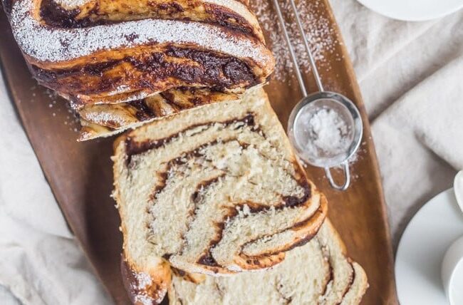 A delicious, twisted loaf of chocolate babka on a wooden platter with powdered sugar sprinkled on top. Slices are placed nearby, echoing the charm of classic apple recipes. Adjacent are a white coffee cup and saucer, a sieve with sugar, and a soft fabric as a backdrop.