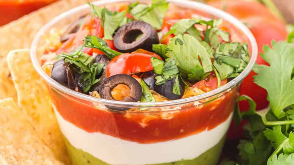 A clear cup filled with layered dip: guacamole at the bottom, followed by sour cream, salsa, and topped with chopped lettuce, diced tomatoes, black olives, and cilantro. Tortilla chips and fresh cilantro surround the cup.