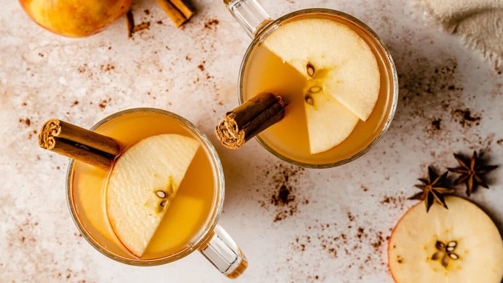 Two clear mugs filled with spiced apple cider, each garnished with a slice of apple and a cinnamon stick. Cinnamon sticks and star anise are scattered on the light-colored surface, along with slices of apple.