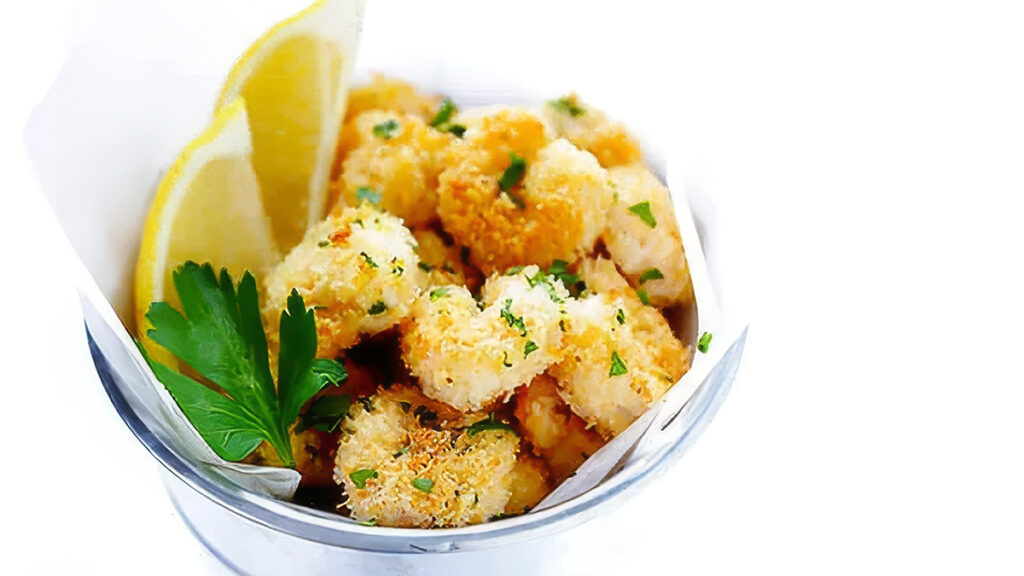 A cup filled with crispy, golden-brown fried shrimp garnished with chopped parsley makes for a delightful entry in finger food recipes. A lemon wedge and a sprig of fresh parsley on the side complete the presentation, all elegantly served in a white paper-lined metal container.