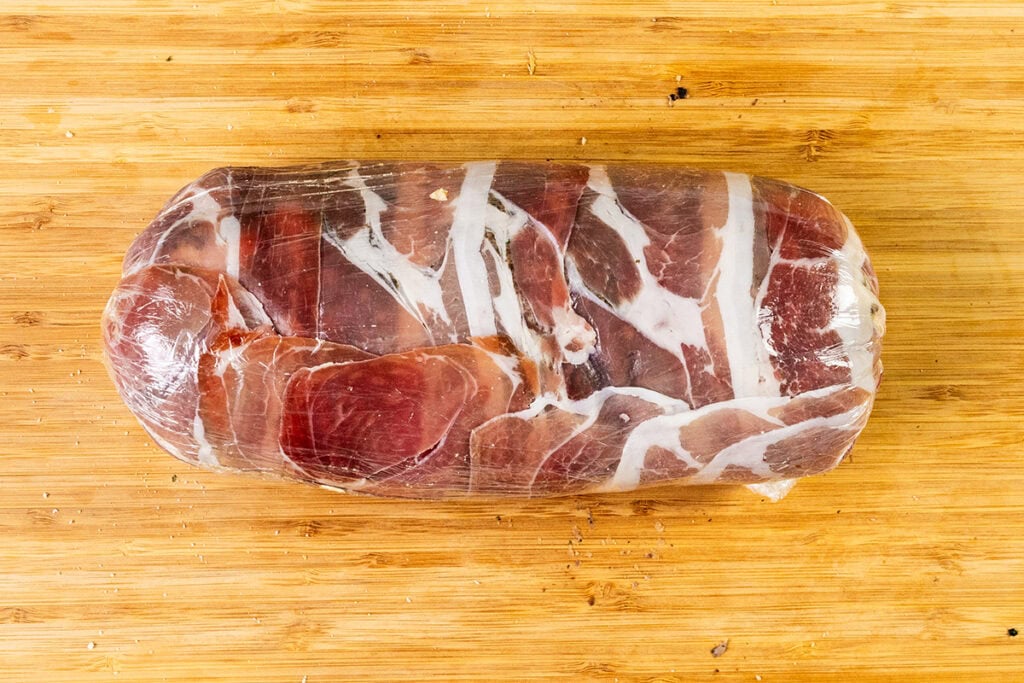 A prosciutto wrapped cut of beef in plastic sits on a wooden cutting board.