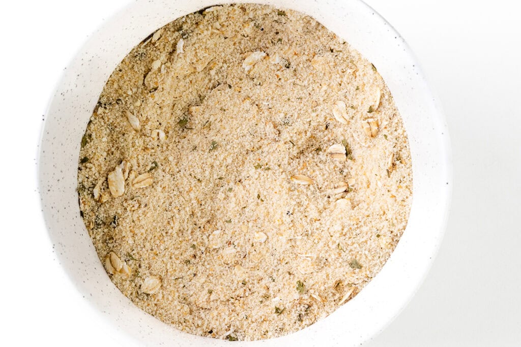 A close-up view of a bowl filled with a mixture of breadcrumbs and herbs, perfect for crafting a delectable Beef Wellington. The blend includes visible oats, adding texture to the breadcrumb mix. The bowl is white with a subtle speckled pattern.