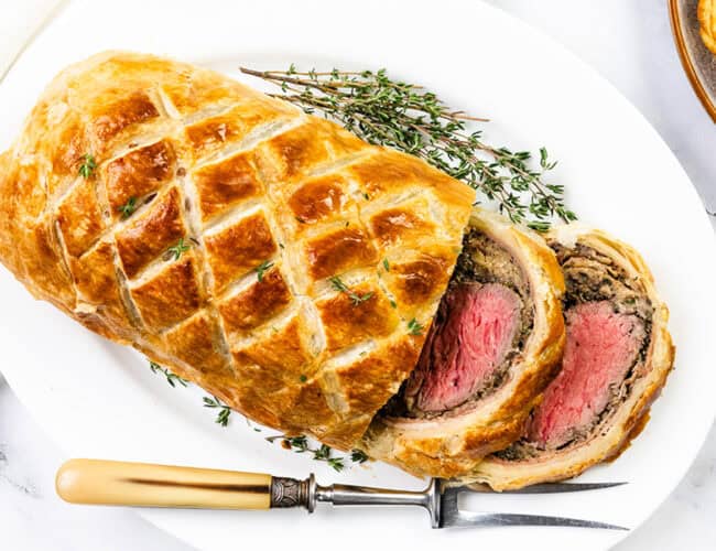 A sliced beef Wellington with golden puff pastry, revealing medium-rare beef and a mushroom duxelles layer, garnished with fresh thyme on a white platter—a festive main dish perfect for Christmas. A vintage fork rests nearby, alongside a white napkin.