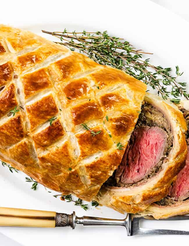 A sliced beef Wellington with golden puff pastry, revealing medium-rare beef and a mushroom duxelles layer, garnished with fresh thyme on a white platter—a festive main dish perfect for Christmas. A vintage fork rests nearby, alongside a white napkin.