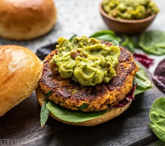 A veggie burger patty on a bun with fresh spinach and a generous scoop of guacamole gives a touch of Cajun-inspired flair. In the background, a bowl of guacamole and a top bun are showcased, surrounded by an assortment of leafy greens.