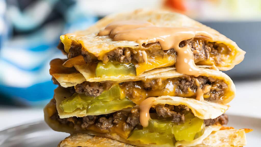 A close-up of stacked cheeseburger quesadillas filled with ground beef, melted cheese, and pickles. A drizzle of sauce tops the layers, showcasing a delicious twist on classic beef recipes against a blurred background.