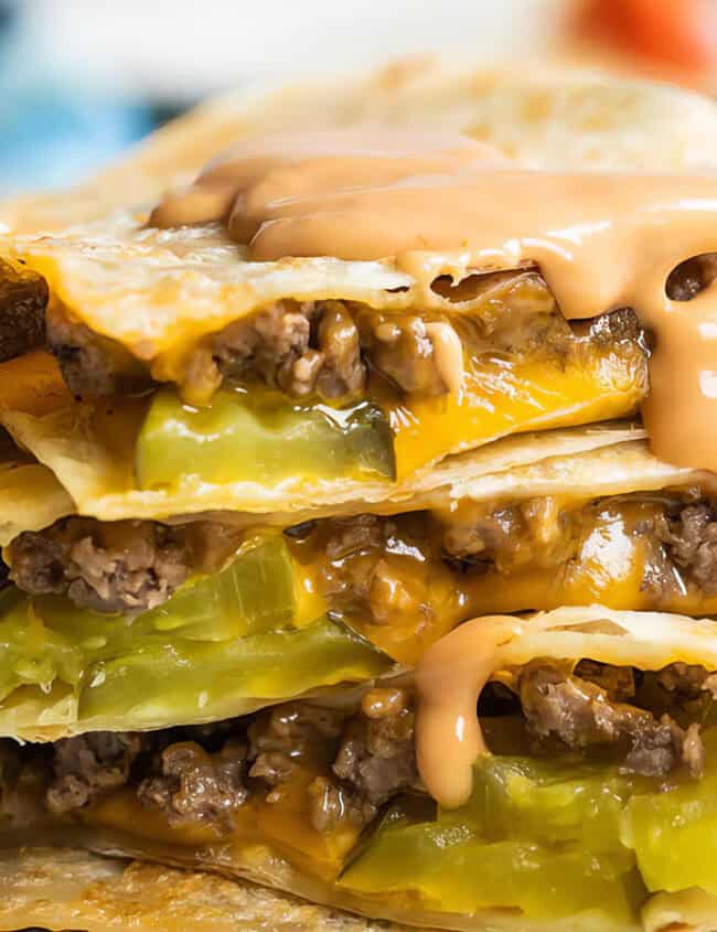 A close-up of stacked cheeseburger quesadillas filled with ground beef, melted cheese, and pickles. A drizzle of sauce tops the layers, showcasing a delicious twist on classic beef recipes against a blurred background.