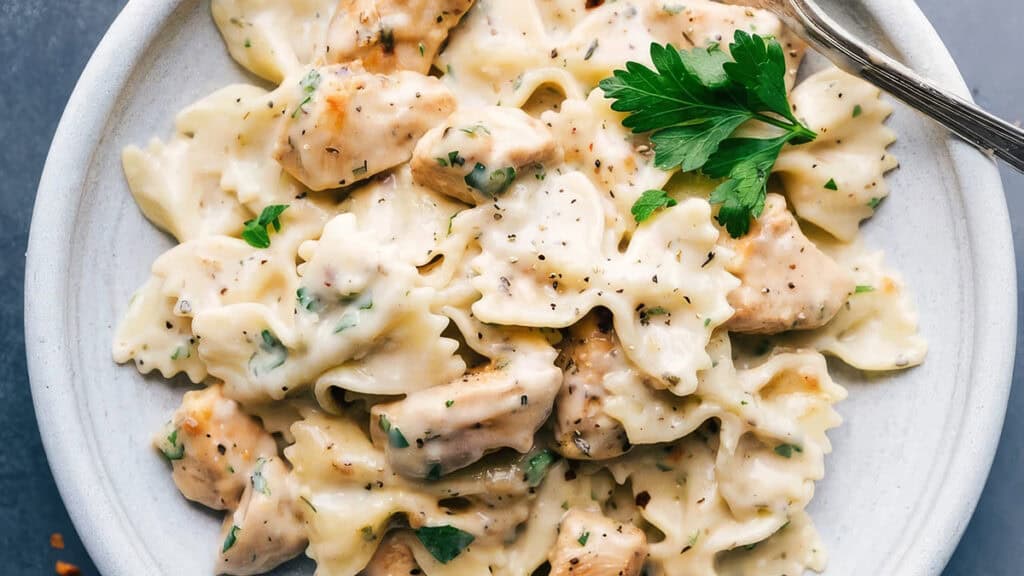 A plate of creamy bowtie pasta with chunks of grilled chicken, garnished with a sprig of parsley.