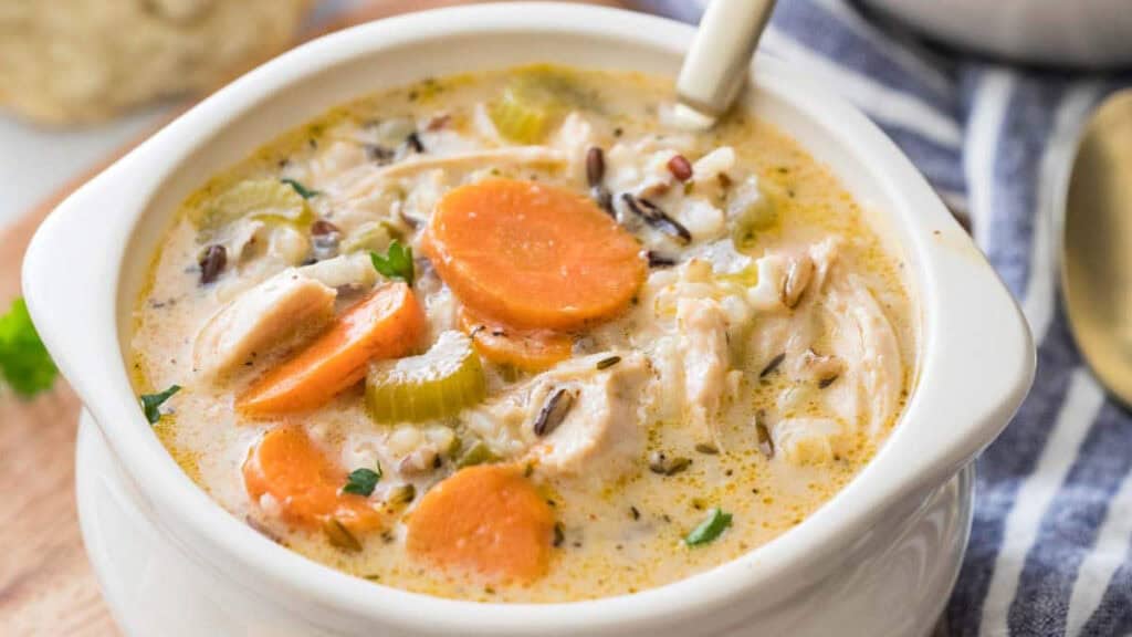 A creamy soup with chunks of chicken, sliced carrots, celery, and wild rice fills a white bowl. A spoon rests inside, inviting you to dive into this one-pot recipe delight. In the background, a striped napkin complements the shine of a gold-tinted spoon.