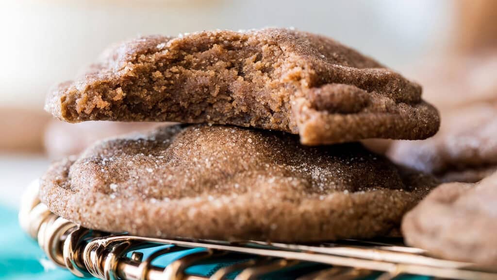 Close-up of a stack of cookies on a cooling rack, perfect for holiday baking. The top cookie has a bite taken out, revealing a soft interior. These brown cookies are sprinkled with sugar for a textured appearance. The softly blurred background adds to the cozy cookie recipes ambience.