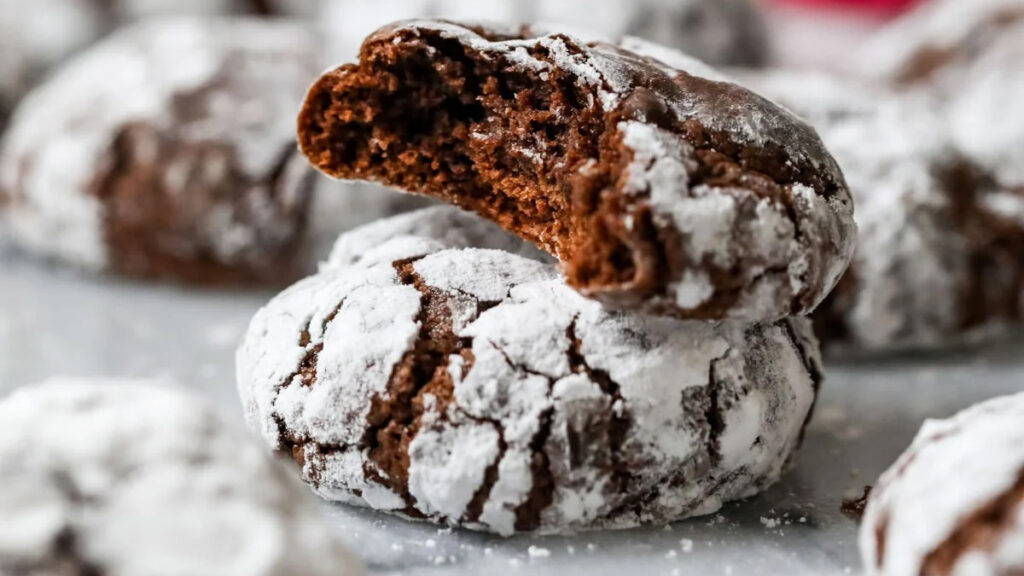 A close-up of two holiday treat chocolate crinkle cookies coated in powdered sugar, one with a bite taken out to reveal a rich, soft interior. The rest of the cookies blur into the background, creating a cozy, inviting scene perfect for festive gatherings.