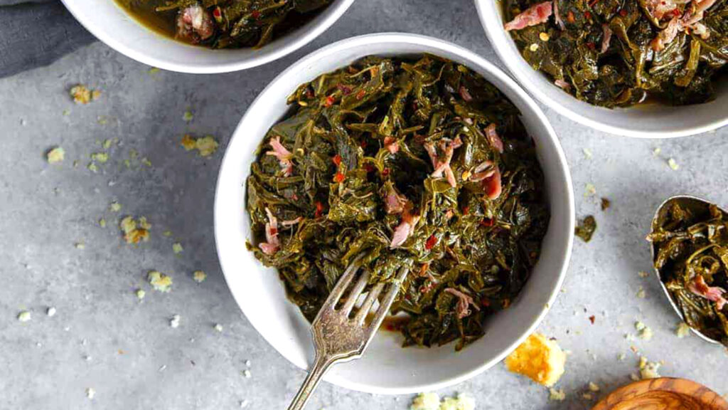 A bowl of cooked collard greens mixed with bits of ham is served in a white dish, embodying the essence of Southern cooking. A fork rests in the bowl, while other similar dishes and crumbs are visible on the gray surface around it.