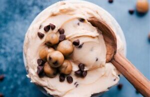 A creamy dessert topped with cookie dough balls and chocolate chips in a jar, inspired by frosting recipes, with a wooden spoon resting on the side. Scattered chocolate chips are visible on the blue surface.