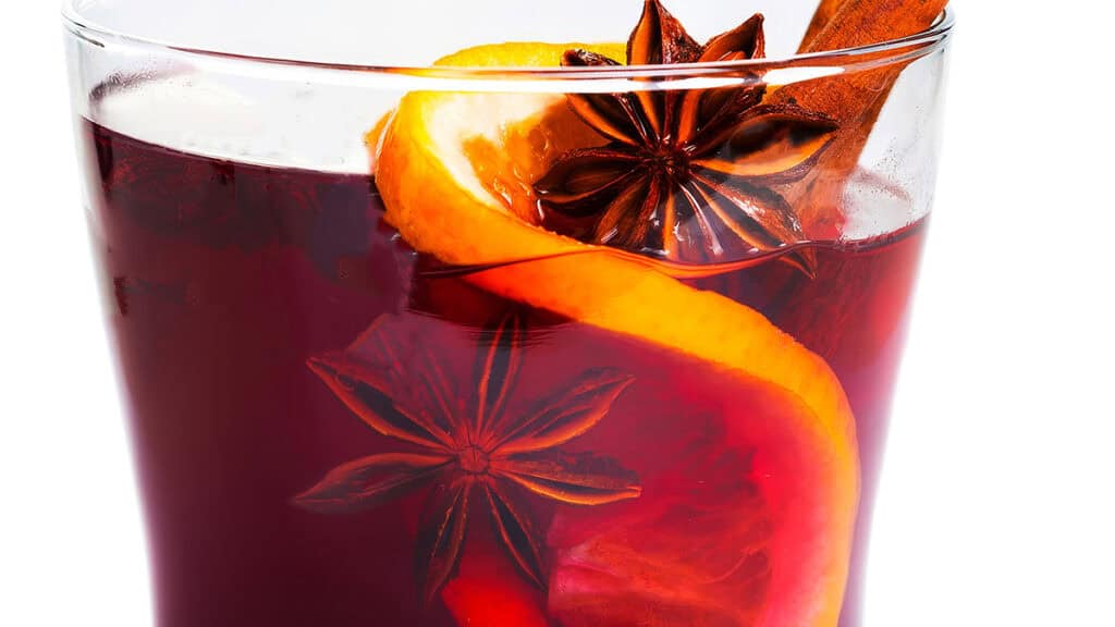 A close-up of a glass filled with mulled wine, featuring a slice of orange, star anise, and a cinnamon stick floating on top. The beverage has a deep red color, and the garnish adds a festive touch.