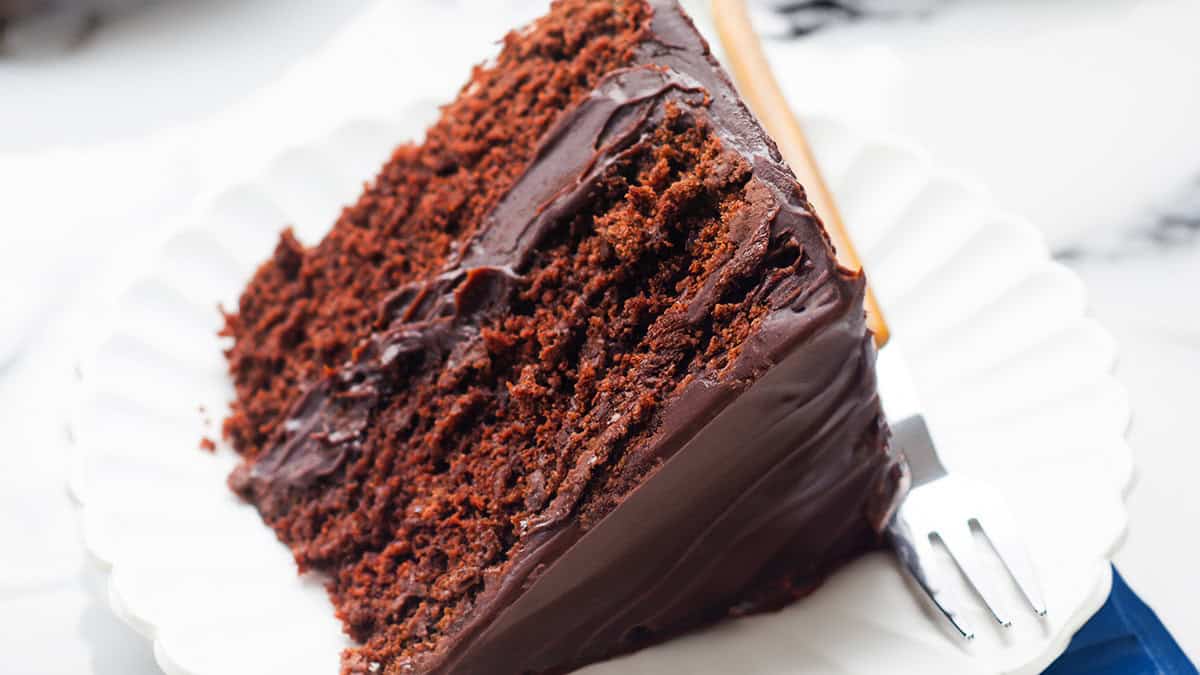 A slice of rich chocolate cake with smooth frosting sits on a white plate, reminiscent of old-fashioned cakes. A fork rests to the side, ready to enjoy the dessert. The background is softly blurred, highlighting the cake's tempting texture and layers.