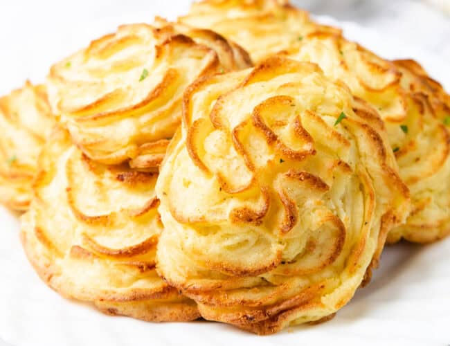 A close-up of golden brown duchess potatoes arranged on a white plate, perfect as festive side dishes. Piped into decorative swirled shapes, they appear crispy and well-baked, adding elegance to any Christmas table.