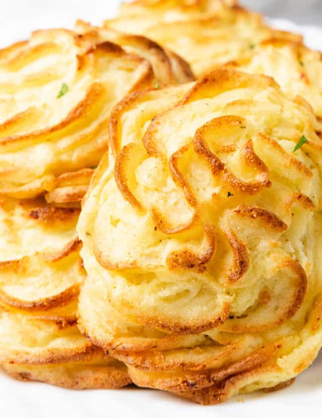 A close-up of golden brown duchess potatoes arranged on a white plate, perfect as festive side dishes. Piped into decorative swirled shapes, they appear crispy and well-baked, adding elegance to any Christmas table.