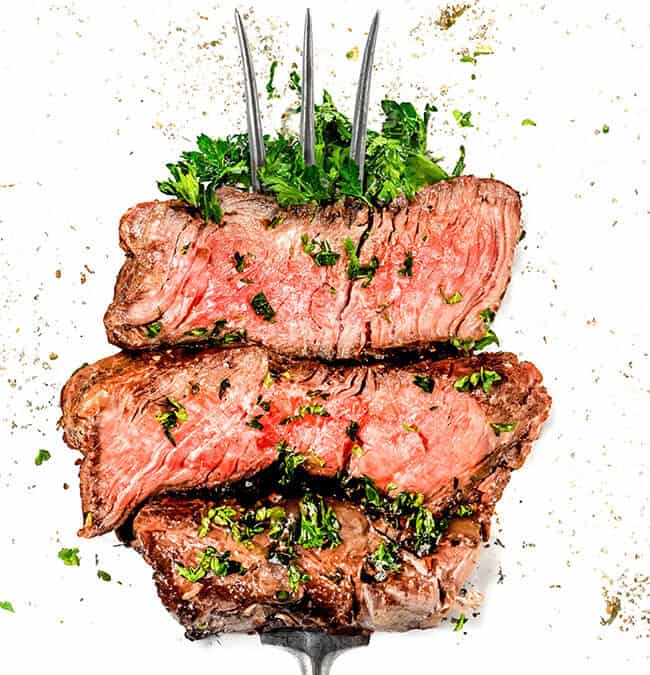 Three slices of medium-rare beef steak are stacked on a fork, garnished with chopped parsley and positioned against a white background sprinkled with black pepper.