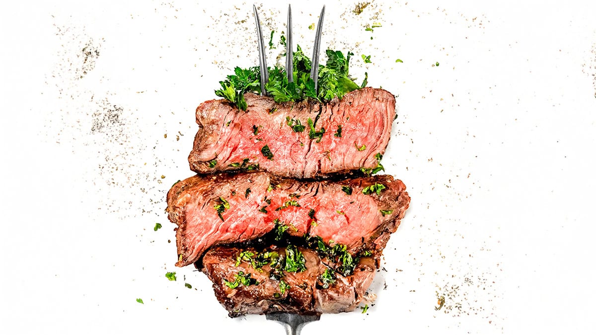 Three slices of medium-rare beef steak are stacked on a fork, garnished with chopped parsley and positioned against a white background sprinkled with black pepper.