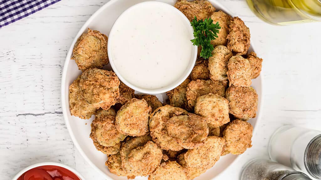 A plate of fried chicken nuggets, emblematic of Southern Cuisine, is served with a bowl of creamy dipping sauce and garnished with parsley. Accompaniments include a small dish of ketchup and bottles of oil and seasonings on a white wooden table.