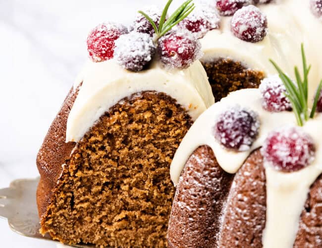 A close-up of a gingerbread Bundt cake topped with white icing, sugared cranberries, and rosemary sprigs is like something straight out of holiday dessert recipes. One slice is cut, revealing the moist interior as it sits on a serving spatula against a white background.