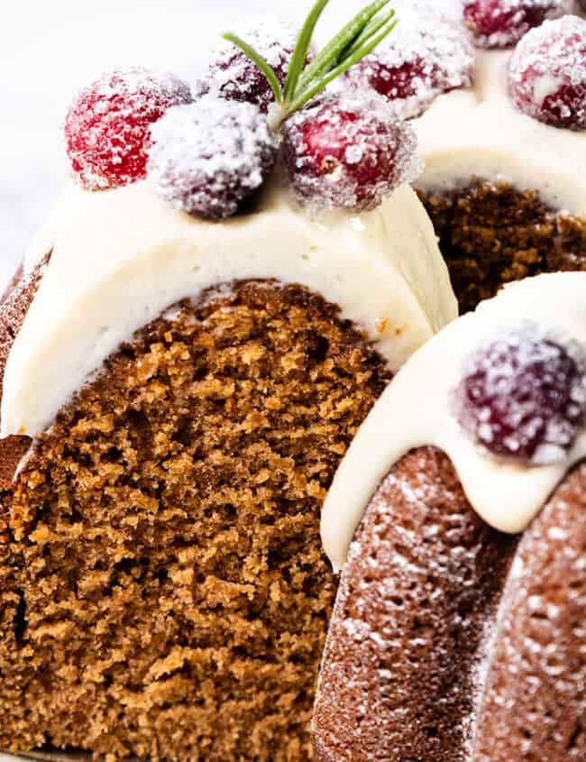 A close-up of a gingerbread Bundt cake topped with white icing, sugared cranberries, and rosemary sprigs is like something straight out of holiday dessert recipes. One slice is cut, revealing the moist interior as it sits on a serving spatula against a white background.