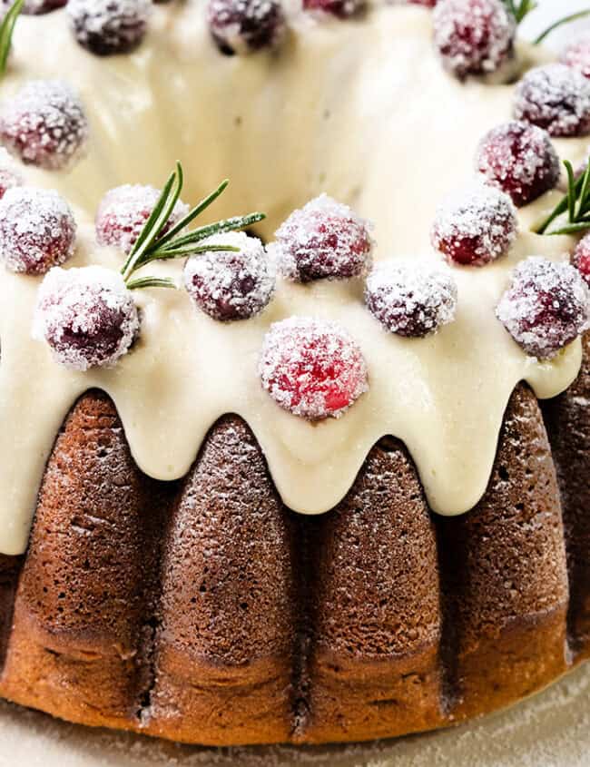 A Christmas bundt cake topped with white icing and garnished with sugared cranberries and sprigs of rosemary on a white plate, perfect for your holiday recipes.