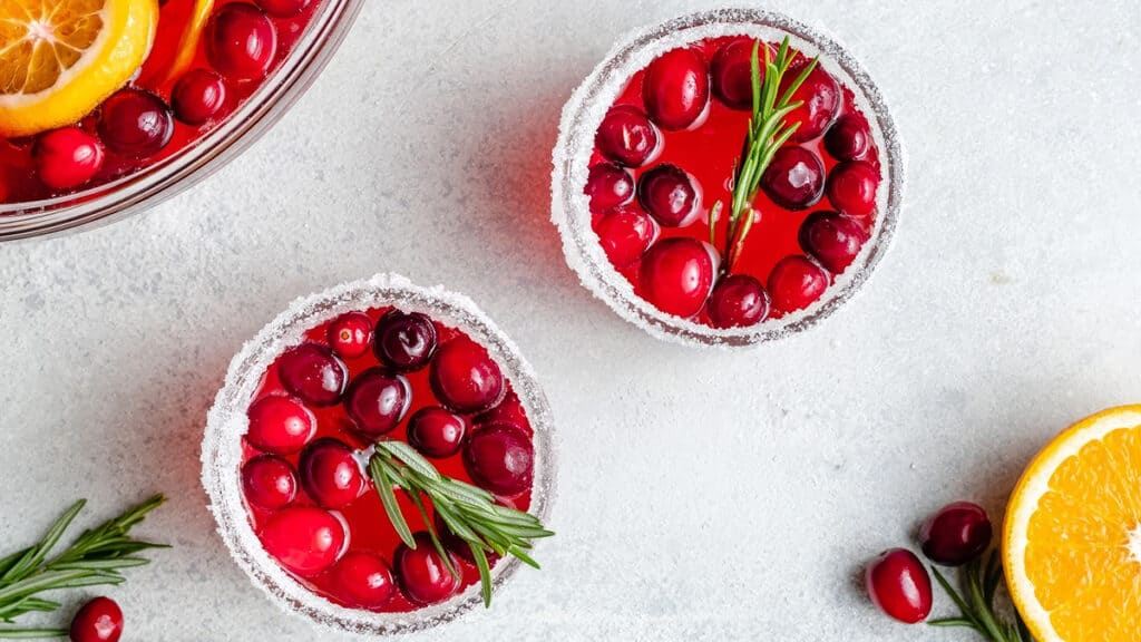 Top view of two glasses filled with cranberry punch, garnished with cranberries and rosemary. The rims are coated with sugar. A sliced orange, cranberries, and rosemary sprigs are nearby on a light gray surface.