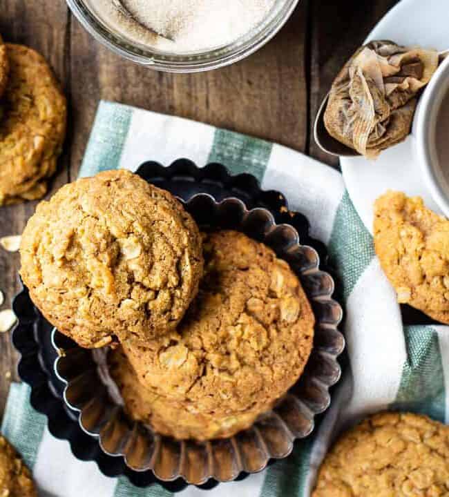 A cozy scene unfolds with homemade oatmeal cookies from cherished cookie recipes nestled in a tin. Scattered oats, a cup of tea with milk on a saucer, and a jar of sugar complete the setting, all serenely laid out on a wooden table adorned with a green striped cloth.