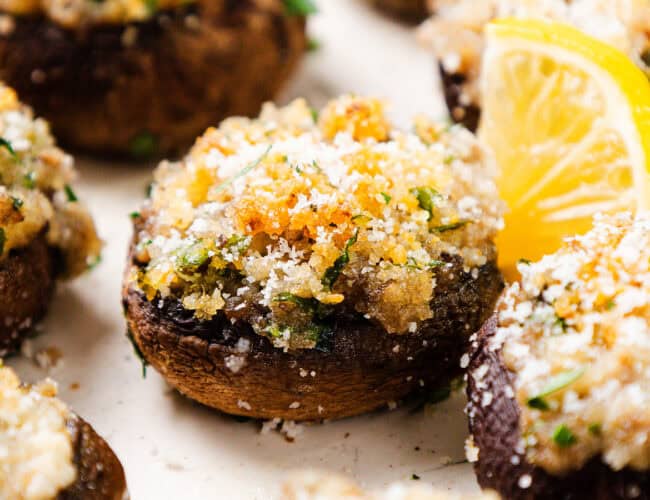 Stuffed mushrooms topped with breadcrumbs and herbs are garnished with parsley and a slice of lemon on the side, making them an ideal party food. Perfect for New Year's festivities, this baked dish boasts a crispy golden-brown crust that pairs wonderfully with various dips.