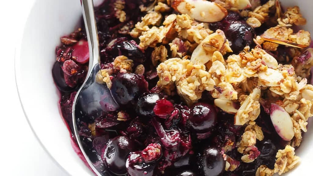 A bowl filled with a blueberry crumble, featuring juicy blueberries and baked oats with almond slices. A spoon rests in the bowl, ready to serve.