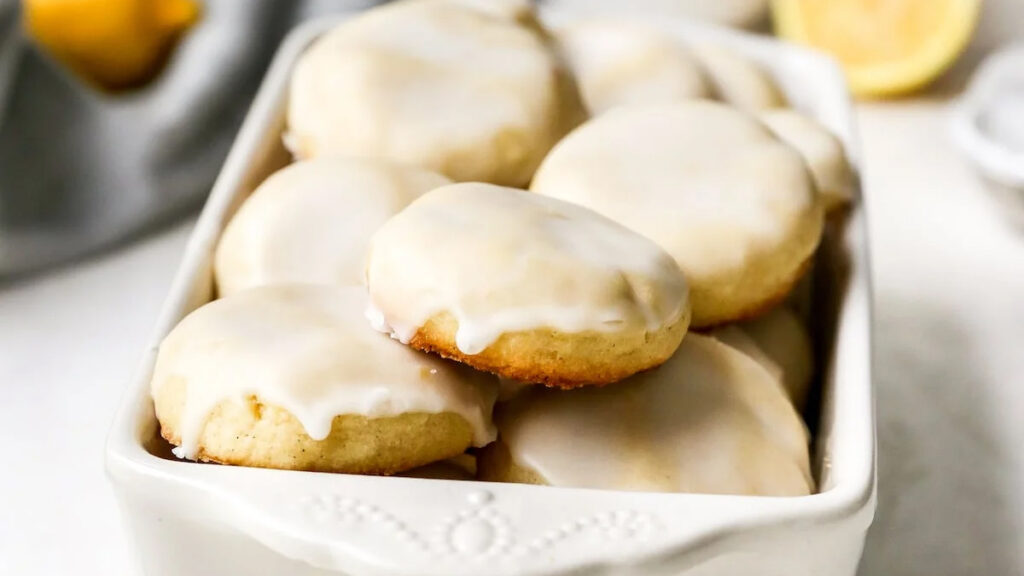 A white ceramic dish filled with round lemon cookies, each topped with a smooth white glaze. These holiday treats are neatly arranged and appear freshly baked, with a golden brown base. A lemon is partly visible in the background.