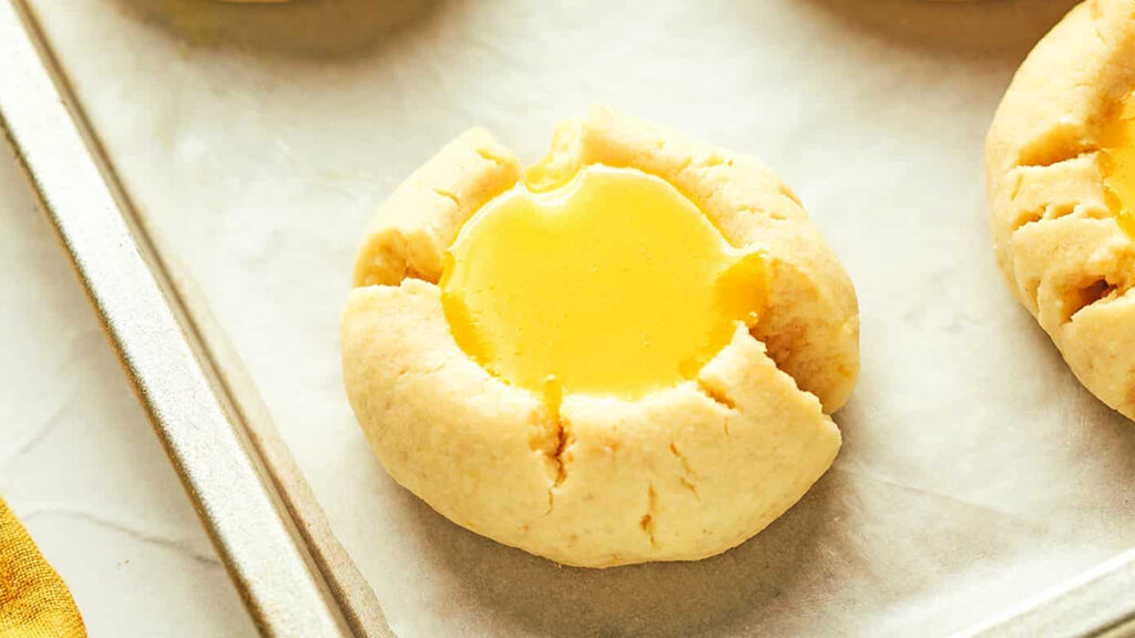 A close-up of a classic lemon thumbprint cookie on a baking sheet lined with parchment paper. The cookie boasts a soft, round shape with vibrant yellow filling at its center, encased in a crumbly, golden brown exterior.